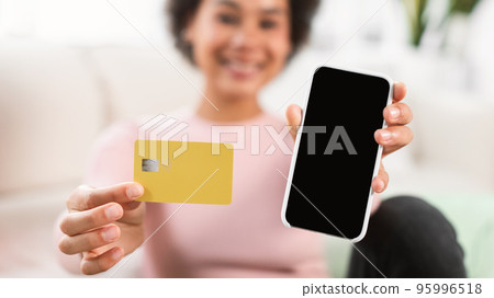 Smiling millennial african american female show credit card and smartphone with blank screen in living room Smiling millennial african american female show credit card and smartphone with blank screen in living room 95996518
