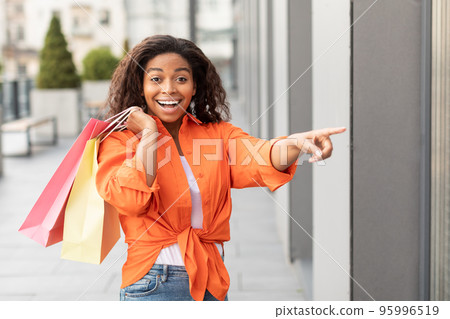 Cheerful surprised emotional funny millennial black woman in casual with a lot of bags points finger at store Cheerful surprised emotional funny millennial black woman in casual with a lot of bags points finger at store 95996519