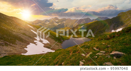 high altitude mountain lake at sunset. beautiful nature landscape of romania. grass and snow on the hills in evening light. sunny weather with clouds on the sky in summer high altitude mountain lake at sunset. beautiful nature landscape of romania. grass and snow on the hills in evening light. sunny weather with clouds on the sky in summer 95997048