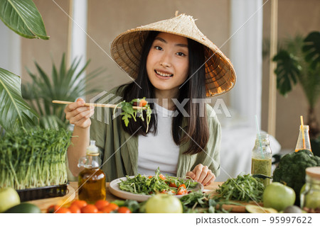 Cheerful young asian woman in traditional conical hat eating healthy salad sitting on the table 95997622