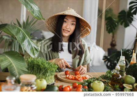 Beautiful asian young woman in traditional conical hat is slicing cherry tomatoes, sitting at table 95997668