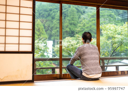 Rear view of a middle-aged man relaxing in a hotel room at a travel destination and looking at the scenery outside 95997747