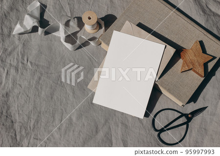 Christmas stationery. Blank greeting card, invitation mockup. Wooden star ornament, old books and white silk ribbon in sunlight. Beige linen tablecloth. Black vintage scissors. Winter wedding flat lay 95997993