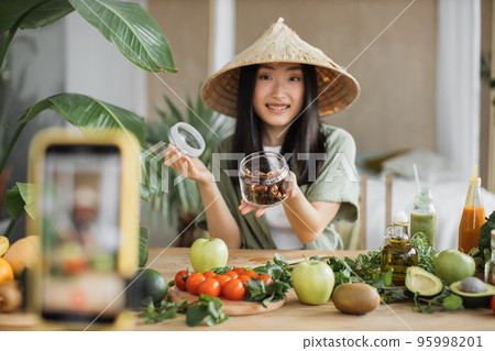 Young woman blogger in traditional conical hat cooking fresh organic healthy food using dates Young woman blogger in traditional conical hat cooking fresh organic healthy food using dates 95998201