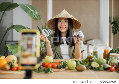 Beautiful smiling asian young woman blogger in traditional conical hat cooking organic healthy food 95998206
