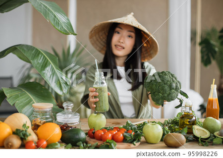 Joyful asian woman in traditional conical hat making smoothies using broccoli Joyful asian woman in traditional conical hat making smoothies using broccoli 95998226