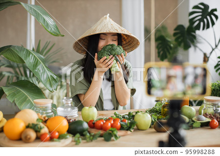 Asian young woman blogger in traditional hat cooking fresh organic healthy food holding broccoli Asian young woman blogger in traditional hat cooking fresh organic healthy food holding broccoli 95998288