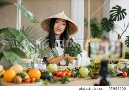 Asian young woman blogger in traditional hat cooking fresh organic healthy food holding broccoli Asian young woman blogger in traditional hat cooking fresh organic healthy food holding broccoli 95998300