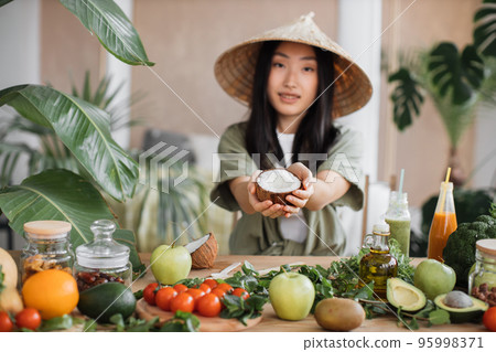 Beautiful asian woman in traditional conical hat holding coconut sitting at table Beautiful asian woman in traditional conical hat holding coconut sitting at table 95998371