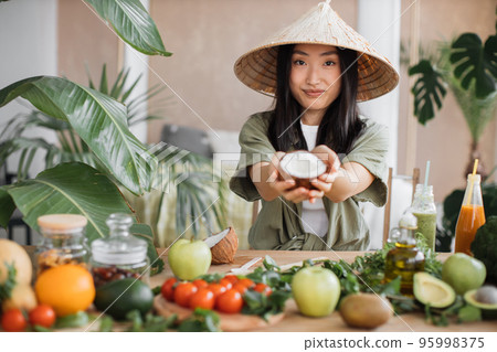 Beautiful asian woman in traditional conical hat holding coconut sitting at table Beautiful asian woman in traditional conical hat holding coconut sitting at table 95998375