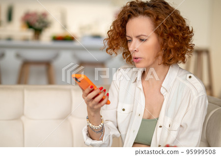 Ginger young woman sitting in the kitchen with a device in hand 95999508