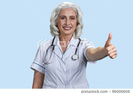 Portrait of aged woman doctor showing thumb up. Smiling senior lady with like gesture. Pale blue background. 96000608