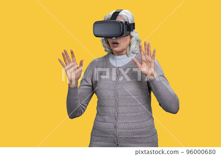 Portrait of astonished senior lady with vr headset. Old woman with her hands up. Isolated on yellow. 96000680