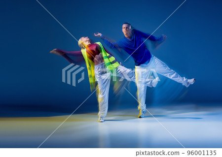 Portrait of young man and woman dancing isolated over dark blue background with mixed lights. Synchronic 96001135