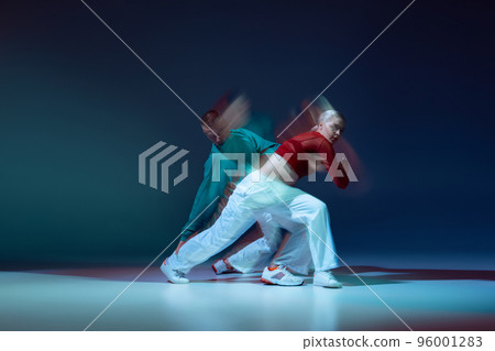 Portrait of young man and woman dancing hip-hop isolated over dark blue background with mixed lights. Training class Portrait of young man and woman dancing hip-hop isolated over dark blue background with mixed lights. Training class 96001283