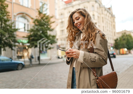 Smiling curly woman wearing warn coat walking down the street and using her phone Smiling curly woman wearing warn coat walking down the street and using her phone 96001679