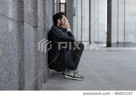 a man in a depressed state sits leaning back against the wall and covering his hands with his palms a man in a depressed state sits leaning back against the wall and covering his hands with his palms 96003187