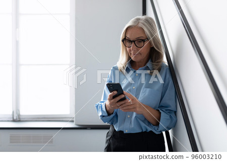 stylish caucasian businesswoman looking at mobile phone while standing against wall in office 96003210