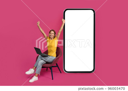 Happy adult european woman sit on chair with laptop, raising hands, celebrate success, victory near huge smartphone 96003470