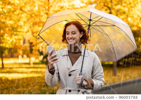 Smiling pretty caucasian millennial female with red hair in raincoat with umbrella enjoys rain, chatting on smartphone 96003788