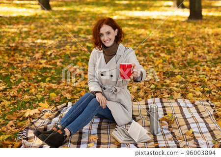 Smiling caucasian millennial female with red hair in raincoat sits on plaid with thermos 96003894
