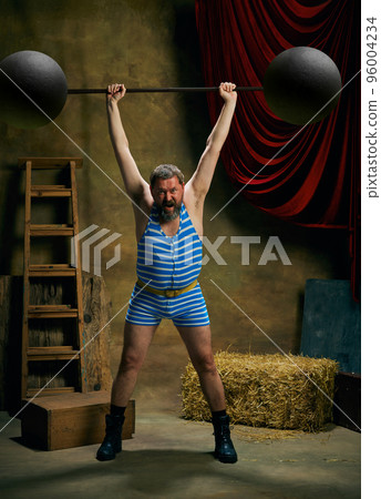 Retro circus. Cinematic portrait of retro circus strongman wearing striped sports swimsuit holding barbell over dark circus backstage background. Art, fashion, style 96004234
