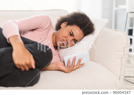 Sad crying millennial african american woman read bad message on phone, lie on sofa, suffers from pain Sad crying millennial african american woman read bad message on phone, lie on sofa, suffers from pain 96004387
