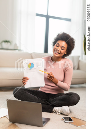 Happy busy millennial african american woman show diagram to laptop webcam sitting on floor in living room 96004488