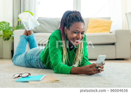 Excited black woman lying on floor with smartphone, working remotely or video chatting at home, free space Excited black woman lying on floor with smartphone, working remotely or video chatting at home, free space 96004636