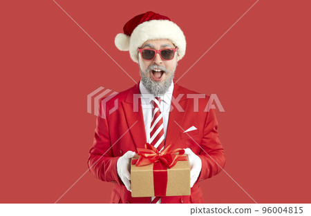 Old grey-haired happy showman in Santa style is giving a present, gift box on red background. 96004815