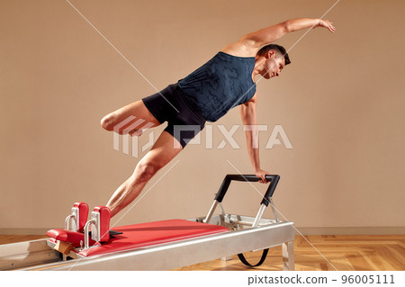 Side view of barefoot male athlete lying on pilates reformer and performing abs exercise during fitness workout. Pilates man concept. Side view of barefoot male athlete lying on pilates reformer and performing abs exercise during fitness workout. Pilates man concept. 96005111