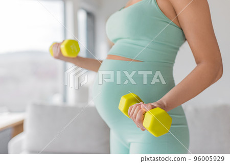 Pregnant woman prenatal training at home doing fitness strength training exercise with dumbbell weights. Body workout arm curls closeup of belly and arms. Easy workout in second and third timester 96005929