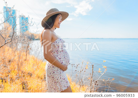 Pregnant Asian woman walking in city park by the lake wearing sun hat during spring nature walk relaxing lookig at landscape. Happy pregnancy outdoor active lifestyle 96005933