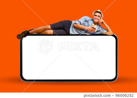 Smiling Young Man Lying On Big Blank Smartphone And Drinking Exotic Cocktail 96006282