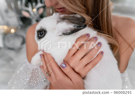 Woman holding a white rabbit symbol of the year 2023. Close-up of a beautiful young blonde woman holding a rabbit in a sparkly dress. She sits in a Christmas decorated room 96008291