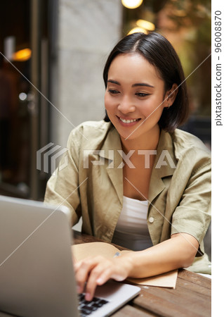 Vertical shot of young asian woman working on remote from outdoor cafe, sitting with laptop and smiling, studying 96008870