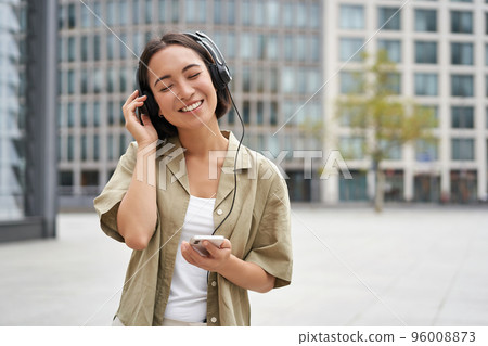 Streaming services concept. Happy asian girl listens music in headphones, holds mobile phone, choosing track or podcast, walking on street 96008873