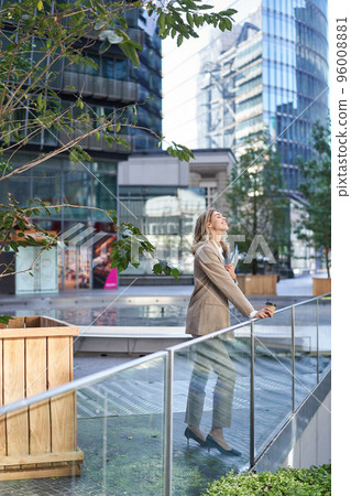 Vertical shot of corporate woman in beige suit standing outside on street, drinks morning coffee takeaway before work 96008881