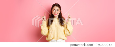 Portrait of beautiful young woman smiling, pointing and looking up at top promo offer, showing advertisement, reading promotional text, standing against pink background Portrait of beautiful young woman smiling, pointing and looking up at top promo offer, showing advertisement, reading promotional text, standing against pink background 96009058