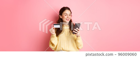 Technology and online shopping. Young pretty lady paying online with credit card, looking at smartphone and smiling, standing over pink background Technology and online shopping. Young pretty lady paying online with credit card, looking at smartphone and smiling, standing over pink background 96009060