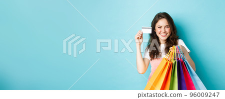 Young satisfied woman smiling, showing plastic credit card and holding shopping bags, buying with contactless payment, standing over blue background Young satisfied woman smiling, showing plastic credit card and holding shopping bags, buying with contactless payment, standing over blue background 96009247