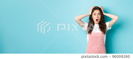 Portrait of troubled and worried young girl, holding hands on head, staring at upper left corner logo banner, standing over blue background 96009260