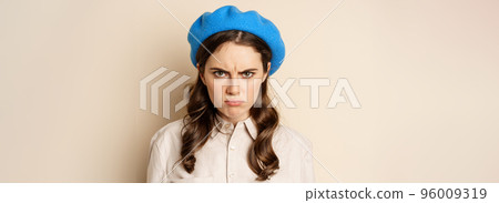 Close up portrait of young beautiful woman sulking, looking angry and disappointed, grimacing displeased, standing over beige background 96009319