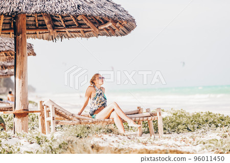 Women sunbathing on dack chair under wooden umbrella on stunning tropical beach. Kiteboarders turquoise blue lagoon of Paje beach, Zanzibar, Tanzania in the background. 96011450