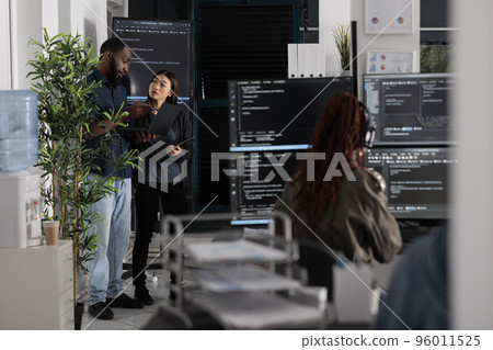 Two app developers analyzing html code looking for errors on laptop computer, checking big tv monitor in it agency office. Coders working on cyber security innovation in busy software company. 96011525
