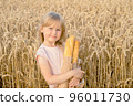 Happy little blonde girl holding a french bread baguette in a wheat field. Children with bread. 96011730