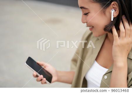 Close up portrait of asian girl, looking at mobile screen, listening music in headphones. Woman with earphones walks on street 96013468