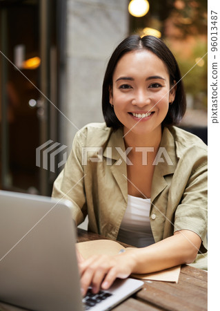 Vertical shot of young asian woman working on remote from outdoor cafe, sitting with laptop and smiling, studying Vertical shot of young asian woman working on remote from outdoor cafe, sitting with laptop and smiling, studying 96013487