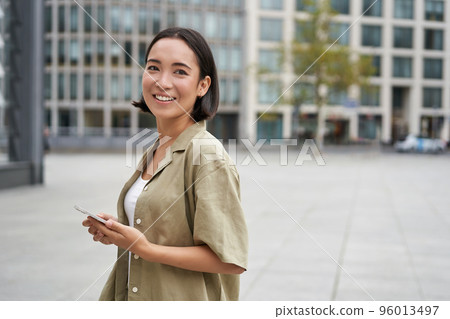 Portrait of asian woman standing on street, city square and holding mobile phone. Girl with smartphone walking outdoors Portrait of asian woman standing on street, city square and holding mobile phone. Girl with smartphone walking outdoors 96013497
