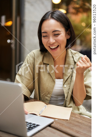 Vertical shot of happy girl talking on video call, looks at laptop, having online meeting, sitting in outdoor cafe Vertical shot of happy girl talking on video call, looks at laptop, having online meeting, sitting in outdoor cafe 96013520